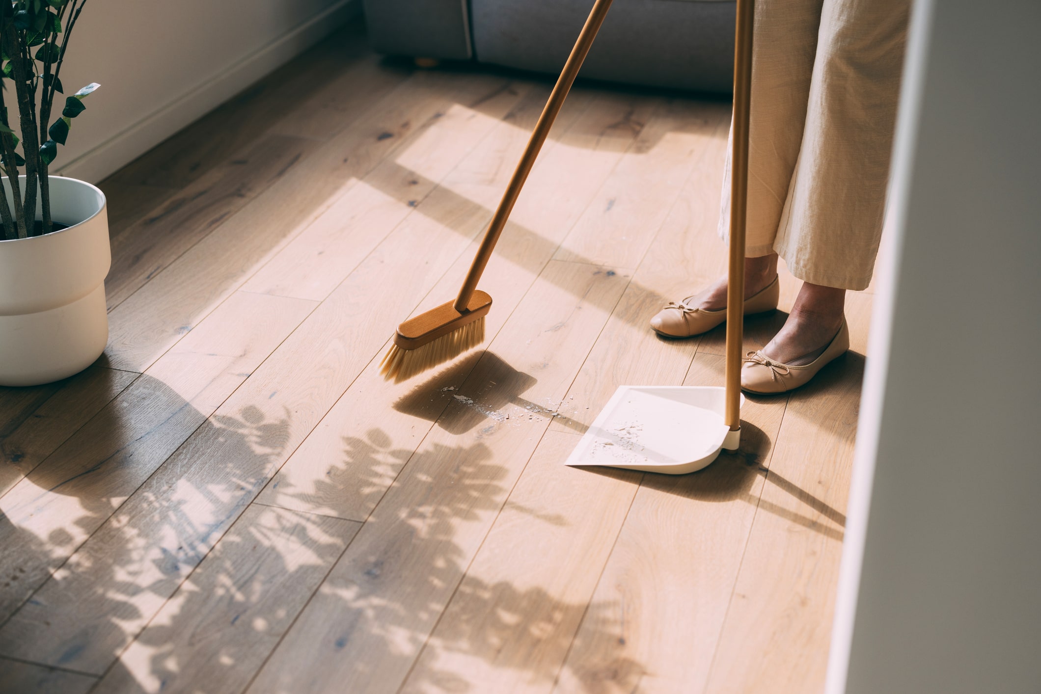 Close-up shot of someone's lower half standing beside a dustpan and broom, sweeping up debris on a sunlit wooden floor indoors.